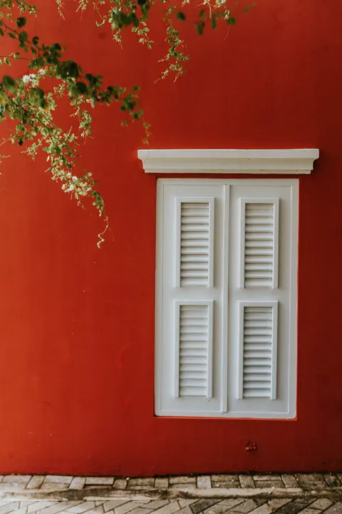 A white shutter set into a deep red Aruba wall, framed by hanging greenery.