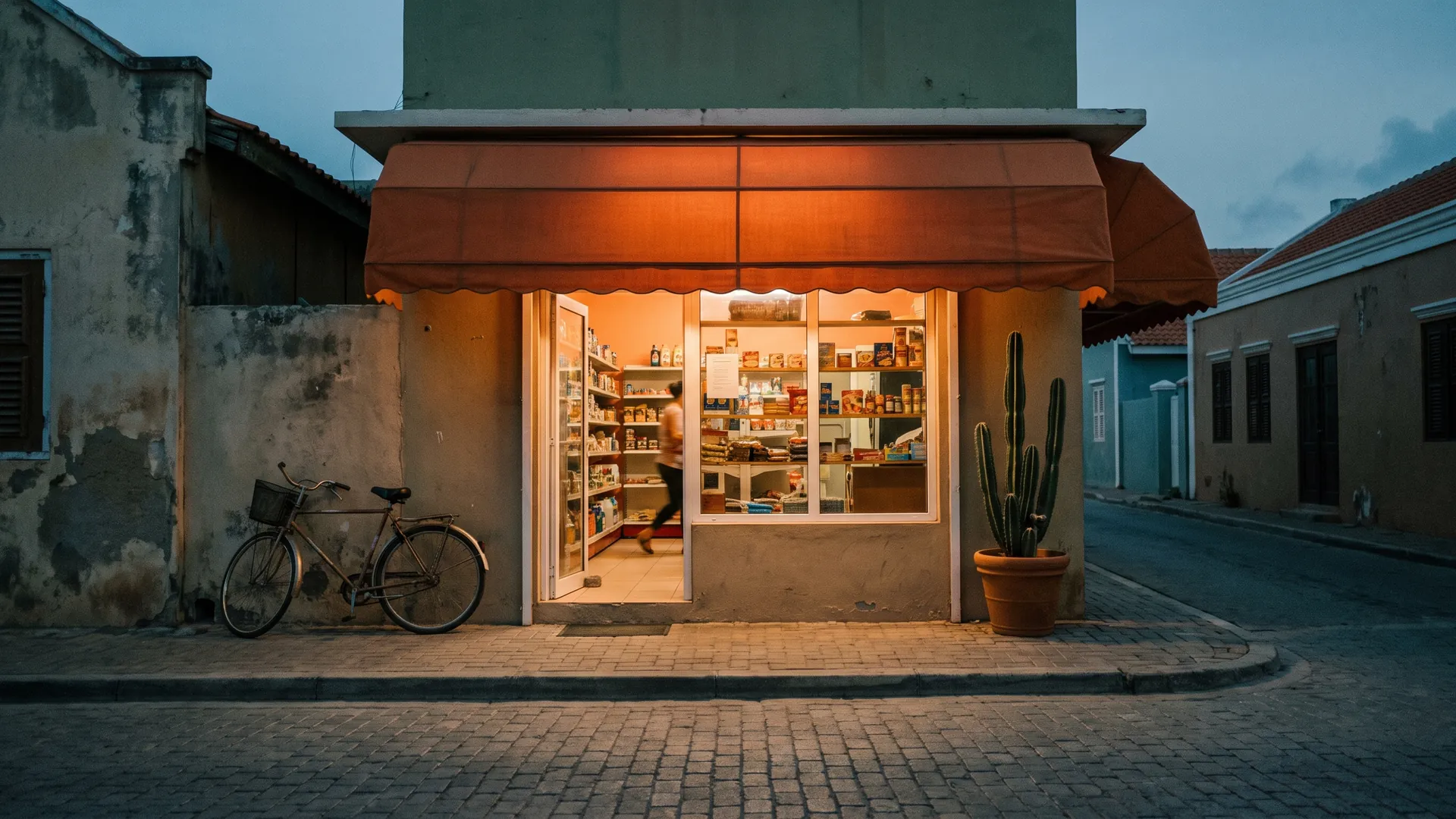 A small corner shop at dusk with an orange awning, its windows lit and warm, a bicycle outside.